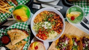 Colorful overhead view of a Mexican food spread, featuring a bowl of rice, beans, grilled chicken, cheese, lettuce, tomatoes, and sour cream, alongside quesadillas, tacos, seasoned fries, and vibrant cocktails garnished with citrus and cherries.